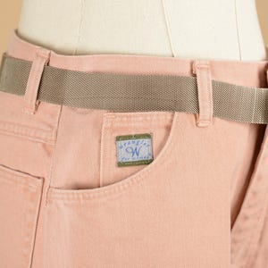 May include: Pink denim pants with a brown woven belt and a Wrangler label on the pocket.