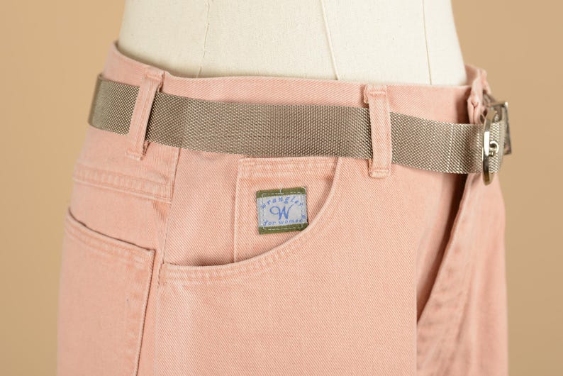 May include: Pink denim pants with a brown woven belt and a Wrangler label on the pocket.