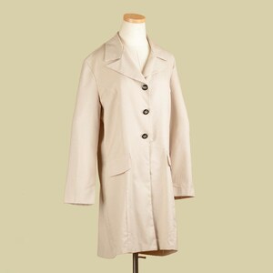 May include: A beige trench coat displayed on a mannequin. The coat has a classic design with a notched collar, three black buttons, and two front pockets. The coat's length extends to the mid-calf.