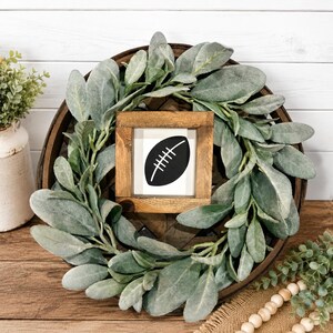May include: A circular wreath made of sage green lamb's ear leaves surrounding a square wooden frame. Inside the frame is a white sign featuring a black graphic of an American football. The wreath is mounted on a dark brown circular wooden base.
