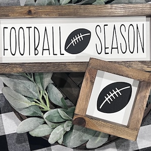 May include: Two rectangular wooden signs with dark brown frames and white centers. The larger sign features the black text FOOTBALL SEASON with a football icon. The smaller square sign features a single black football icon in the center.
