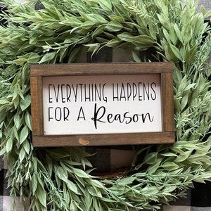 May include: A wooden sign with the text "Everything Happens For A Reason" in black lettering on a white background. The sign is framed in a dark brown wood frame and is surrounded by a green leafy wreath.