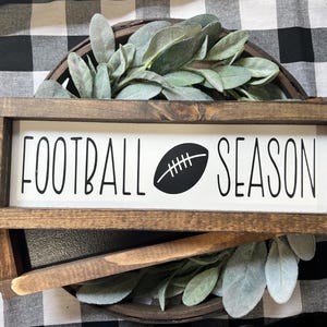 May include: A rectangular wooden sign with a dark brown frame and a white center panel. The text "FOOTBALL SEASON" is printed in black, with a black football graphic replacing the letter "O" in the word "FOOTBALL" and appearing between the words.