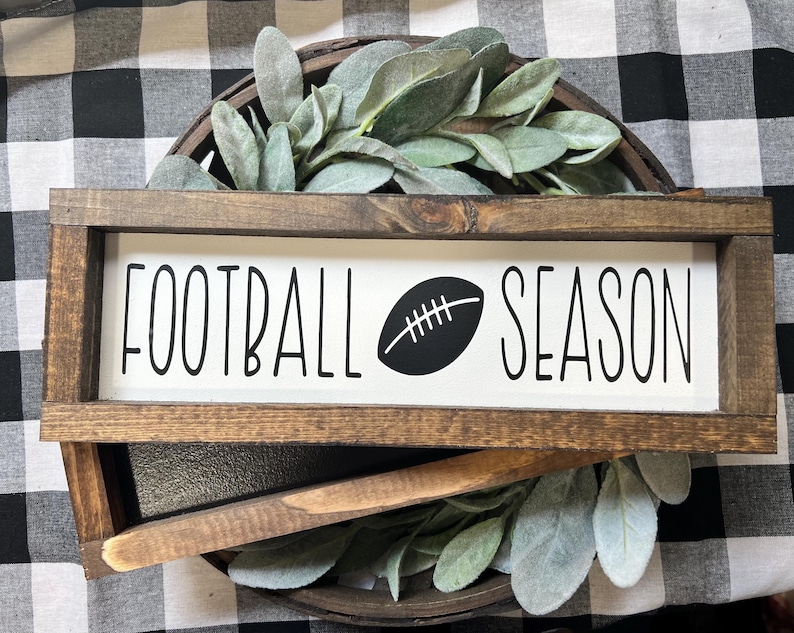 May include: A rectangular wooden sign with a dark brown frame and a white center panel. The text "FOOTBALL SEASON" is printed in black, with a black football graphic replacing the letter "O" in the word "FOOTBALL" and appearing between the words.