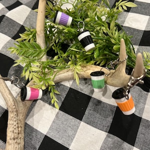 May include: Miniature coffee cup keychains in various colors, including purple, black, pink, green, and orange. Each keychain has a silver clasp. The keychains are arranged on a decorative background with greenery and antlers.