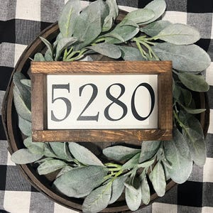 May include: A wooden sign with a white background and black numbers. The sign reads "5280". The sign is framed in a dark brown wood frame.