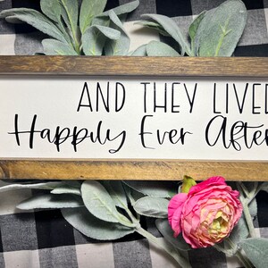 May include: A wooden sign with a white background and black text that reads "And they lived happily ever after". The sign is framed in a dark brown wood frame.