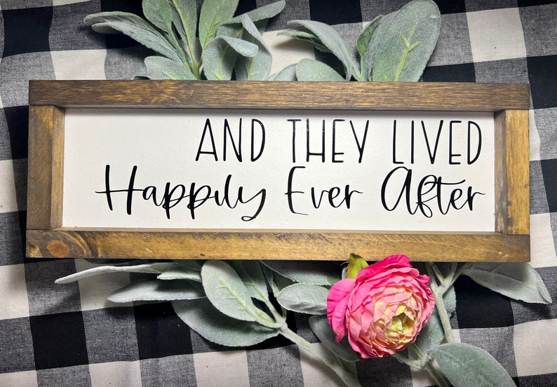 May include: A wooden sign with a white background and black text that reads "And they lived happily ever after". The sign is framed in a dark brown wood frame.