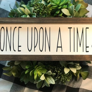 May include: A wooden sign with the text "ONCE UPON A TIME" in black lettering and a small black heart on the right side. The sign is framed in a dark brown wood frame.