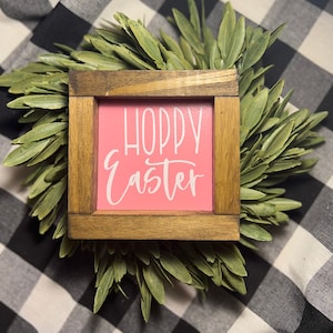 May include: A small wooden sign with a pink background and white lettering that says "Hoppy Easter". The sign is surrounded by green leaves.