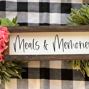 May include: A wooden sign with the text "Meals & Memories" in black script font. The sign is on a black and white checkered background with green foliage on either side.