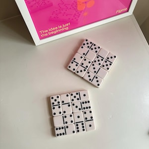 May include: Two sets of off-white dominoes with black pips arranged in square formations. Above them sits a white-framed print featuring a bright pink background with the text "The idea is just the beginning" and "Figma" in yellow.