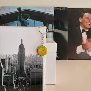 May include: A silver key chain with a yellow and green Topo Chico bottle cap charm. The charm has the words "Twist of Lime" and "Topo Chico" on it. The keychain is hanging from a white surface.