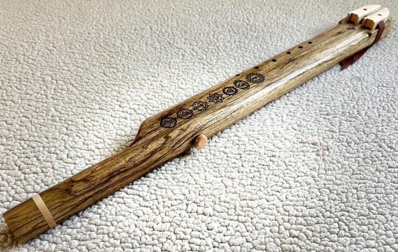 May include: A wooden flute with a brown leather strap and a series of symbols carved into the wood. The flute is approximately 24 inches long.