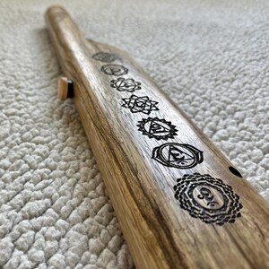 May include: A wooden flute with seven chakra symbols engraved on it. The symbols are in black and white and are arranged in a row along the length of the flute.