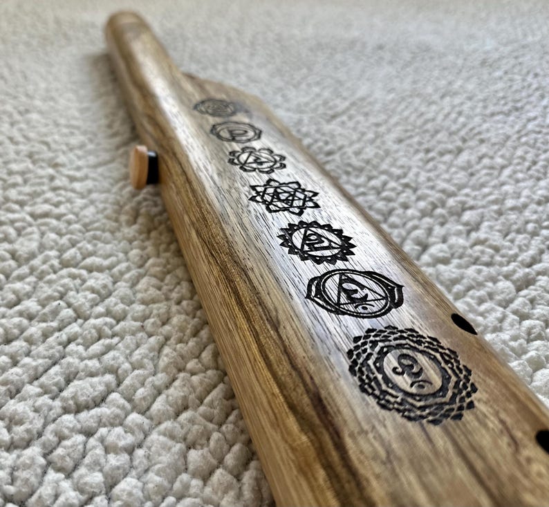 May include: A wooden flute with seven chakra symbols engraved on it. The symbols are in black and white and are arranged in a row along the length of the flute.