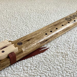 May include: A wooden flute with a brown and black wood grain pattern. The flute has a mouthpiece and a series of holes along the top. The flute is decorated with a series of small circles and a leather strap.