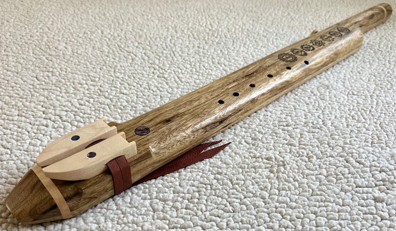 May include: A wooden flute with a brown and black wood grain pattern. The flute has a mouthpiece and a series of holes along the top. The flute is decorated with a series of small circles and a leather strap.