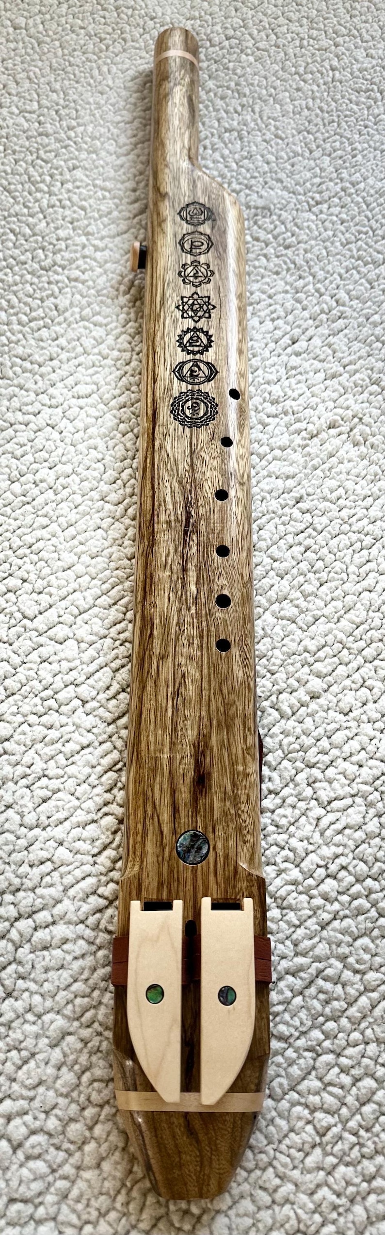 May include: A wooden flute with seven holes and a decorative design of seven chakras. The flute is made of dark wood with a light wood inlay at the bottom. The flute is decorated with a mother of pearl inlay.