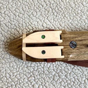 May include: A wooden flute with two decorative inlays made of abalone shell. The flute is brown with a light brown wood trim. The inlays are green and iridescent.