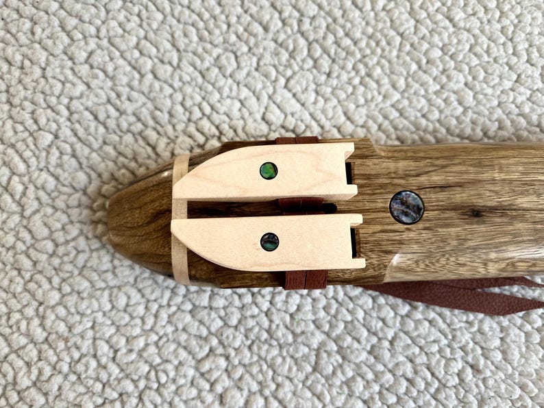 May include: A wooden flute with two decorative inlays made of abalone shell. The flute is brown with a light brown wood trim. The inlays are green and iridescent.