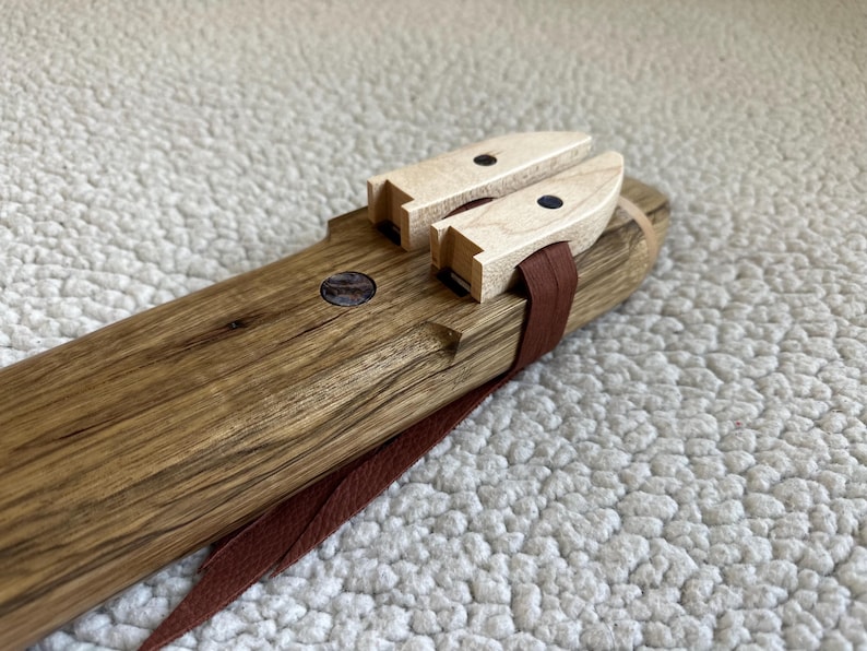 May include: A wooden flute with a brown leather strap and two wooden tuning keys with black dots. The flute is made of dark wood with a natural grain.