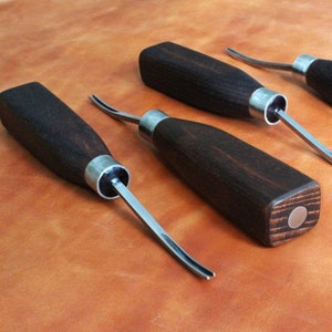 Può includere: Quattro strumenti per intagliare il cuoio con manici in legno e lame in metallo. Gli strumenti hanno manici di colore marrone scuro e dettagli in metallo argentato.