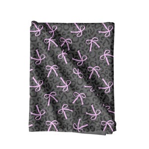 May include: A folded textile featuring a grey leopard print with a repeating pattern of pink bows. The bows are evenly distributed across the fabric. The material appears soft and could be used for various craft projects or as a decorative item.