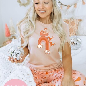 May include: A pink t-shirt with a graphic of Santa Claus riding a rainbow with reindeer. The t-shirt is worn by a person sitting on a white bed with a white blanket. The text "Digital download PNG" is on a pink circle in the bottom left corner of the image.