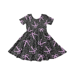 May include: A short-sleeved dress with a dark grey leopard print and light pink bows. The dress has a round neckline and a flared skirt, suitable for casual wear.