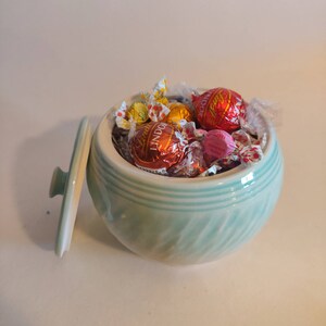 May include: A light blue ceramic bowl with a lid, filled with assorted chocolate candies wrapped in foil. The candies are red, pink, orange, and yellow.