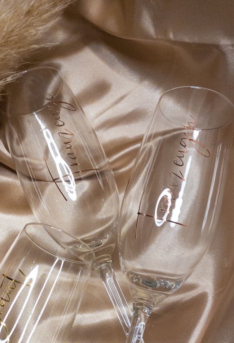 May include: Three champagne flutes with gold lettering. The flutes are on a beige fabric background. The lettering on the flutes reads "Forever", "Always", and "Together".