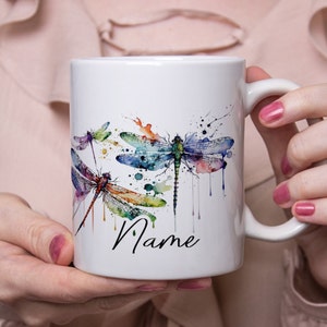 Könnte beinhalten: Weiße Keramiktasse mit einem Aquarelldesign von drei bunten Libellen und dem Wort "Name" auf der Vorderseite.