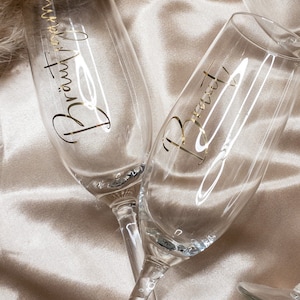 May include: Two champagne flutes with gold lettering. One flute says "Bride" and the other says "Bridesmaid". The flutes are on a light brown satin fabric.