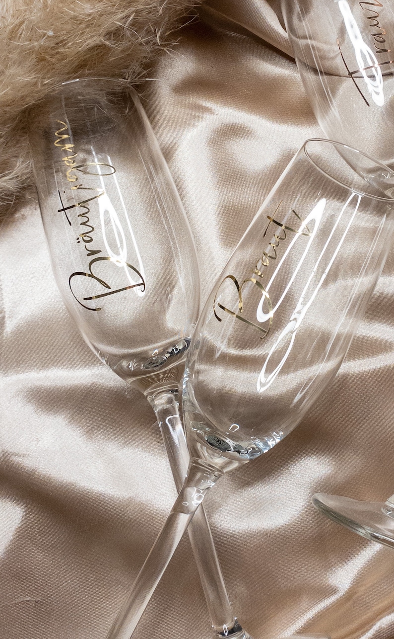 May include: Two champagne flutes with gold lettering. One flute says "Bride" and the other says "Bridesmaid". The flutes are on a light brown satin fabric.