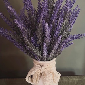 May include: A bouquet of lavender flowers in a white vase. The lavender has purple blooms and green stems. The vase is tied with a cream-colored lace ribbon. The background is a muted gray.