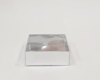 May include: A small, square silver box with a clear, shiny lid. The box is sitting on a white surface. The lid appears to be sealed with clear tape. The box is likely for packaging small items.