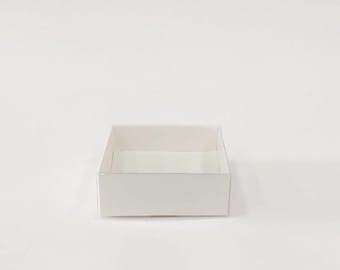 May include: A small, square, white cardboard box with a shallow depth. The box is empty and sits on a white surface. The box has a simple, clean design.