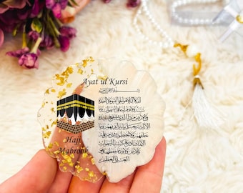 May include: A decorative, scalloped-edge resin piece with gold flakes, featuring Arabic script and an illustration of the Kaaba. Text includes "Ayat ul Kursi" and "Hajj Mabrook". A white pearl prayer bead necklace and flowers are in the background.
