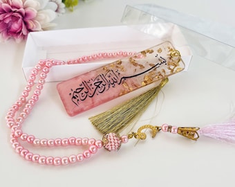 May include: A pink beaded prayer bead set with a matching bookmark. The bookmark is pink with gold leaf accents and Arabic calligraphy. The set includes a gold tassel and a pink tassel.