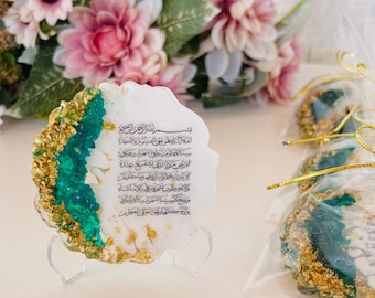 May include: A white, round resin art piece with a green and gold design. The piece features an Arabic inscription in black ink. The inscription reads "Bismillah ir-Rahman ir-Rahim, Allahumma inni as'aluka al-shifa'a, wa as'aluka al-'afiya, wa as'aluka min sharri ma khalaqta". The piece is on a clear acrylic stand.