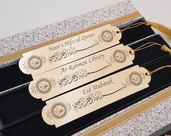 May include: Gold-colored metal bookmarks with Arabic calligraphy and decorative tassels. The bookmarks are in a gift box with a patterned design. The text on the bookmarks includes "Noor's Hifz-ul Quran", "Ar-Rahman Library", and "Eid Mubarak".