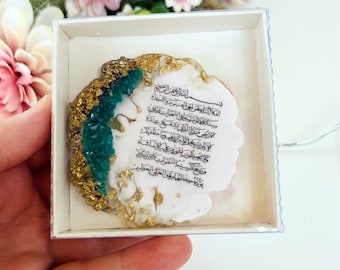 May include: A white circular resin art piece with gold and green accents. The art piece features Arabic calligraphy in black ink. The art piece is displayed in a white box with a clear plastic lid.