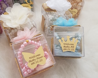 May include: A collection of small, clear boxes with gold trim, each containing a pearl necklace and a decorative flower. The boxes have personalized labels with the text "Welcome Baby Noor 09.11.22" and "Welcome Baby Ali 09.11.22".