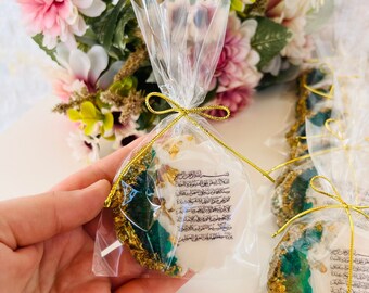 May include: A clear cellophane bag tied with gold ribbon contains a white, irregularly shaped item with gold leaf accents and green and gold edges. The item has Arabic text printed on it. A bouquet of flowers is in the background.
