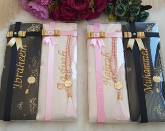 May include: Four gift boxes with pink and gold ribbon bows. Each box contains a white prayer rug with a gold tassel and a pink beaded prayer chain. The boxes are labeled with the names 'Ibrahim', 'Aisha', 'Hafsah', and 'Muhammad'.
