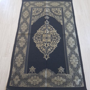 May include: A black prayer rug with gold embroidered floral patterns. The rug has a central design with a repeating pattern of flowers and leaves. The rug is bordered by a decorative pattern.