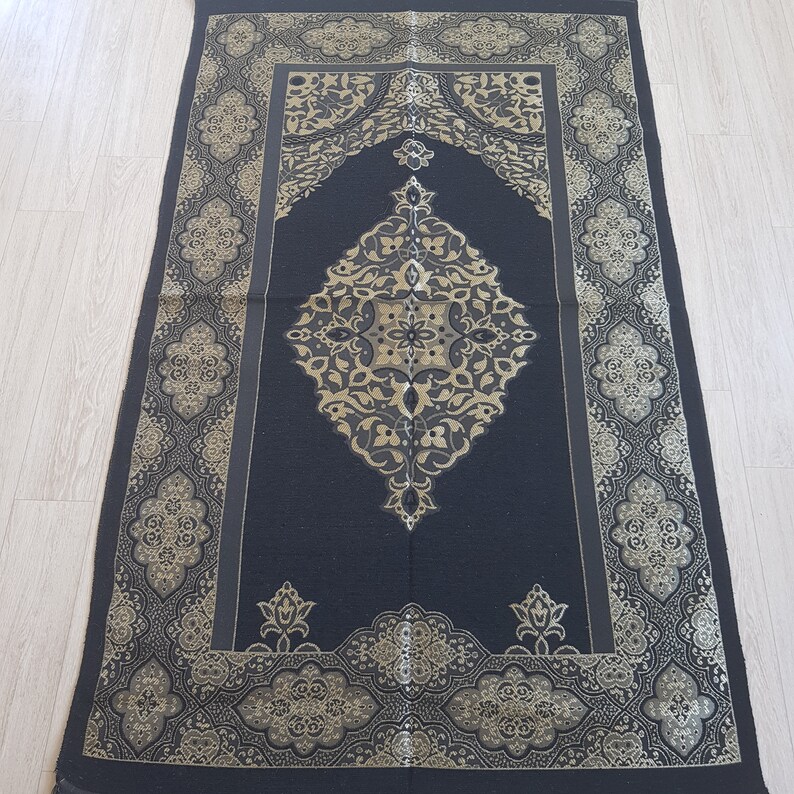 May include: A black prayer rug with gold embroidered floral patterns. The rug has a central design with a repeating pattern of flowers and leaves. The rug is bordered by a decorative pattern.