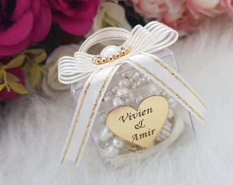 May include: A clear plastic box with a white ribbon and a gold heart charm. The heart charm has the names "Vivien & Amir" engraved on it. The box contains a white pearl necklace.