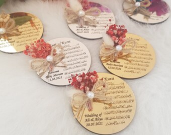 May include: A set of five round, gold-colored, mirrored acrylic plaques with Arabic calligraphy and text in English. The plaques are decorated with dried flowers and a pearl. The text on the plaques includes "Welcome Baby Mariam", "Wedding of Ali & Aliya", and "Ayat ul Kursi".